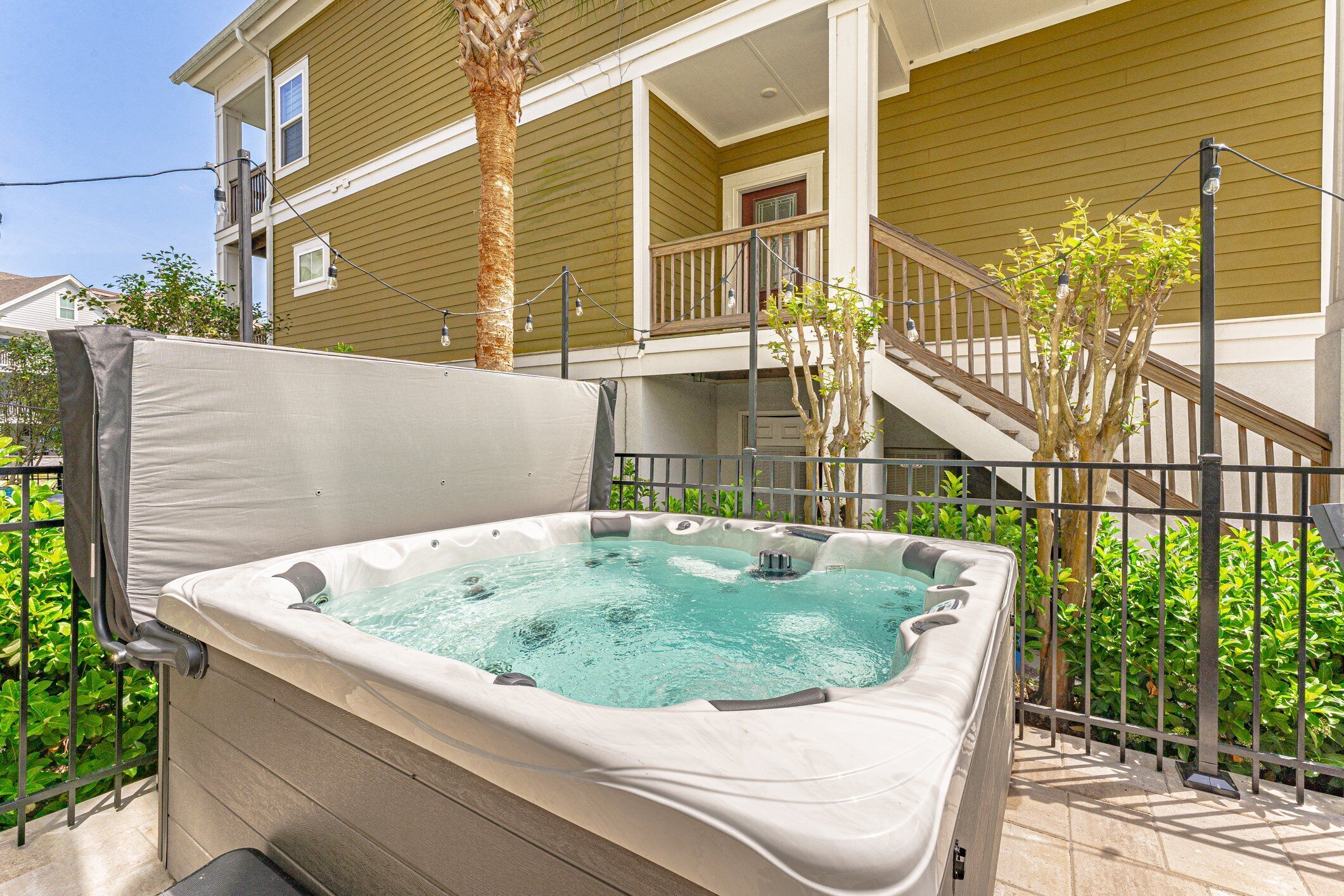 A hot tub in a backyard with a wooden fence, a palm tree, and a house in the background.