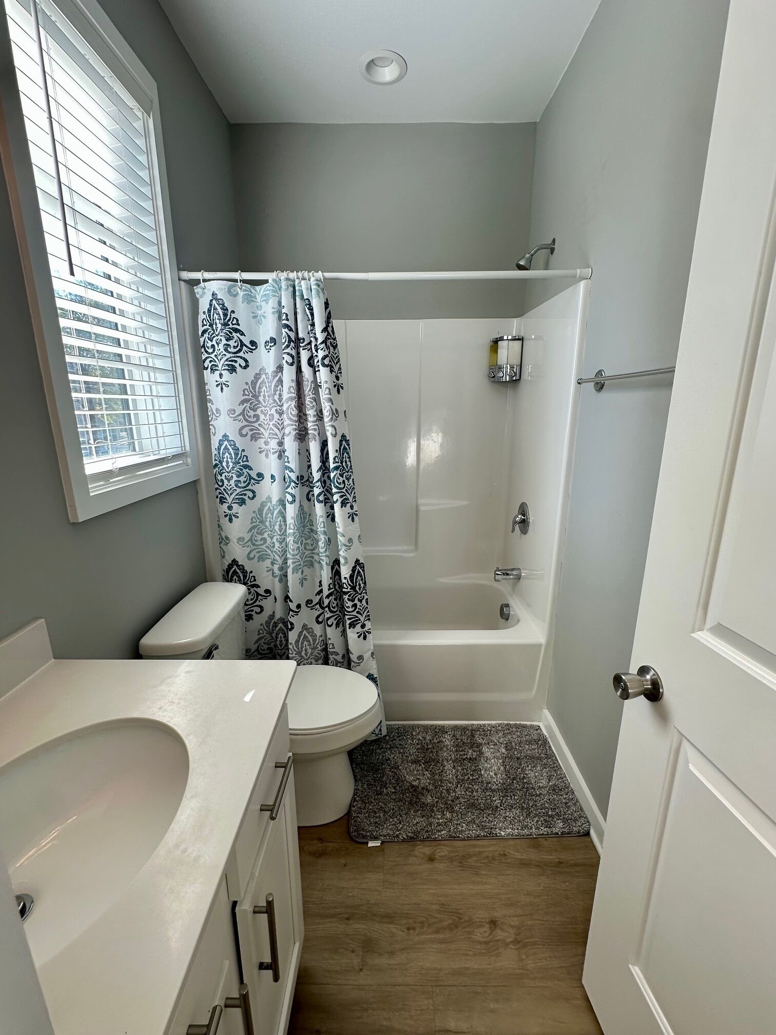 A bathroom with a toilet, sink, shower, and a window with blinds.