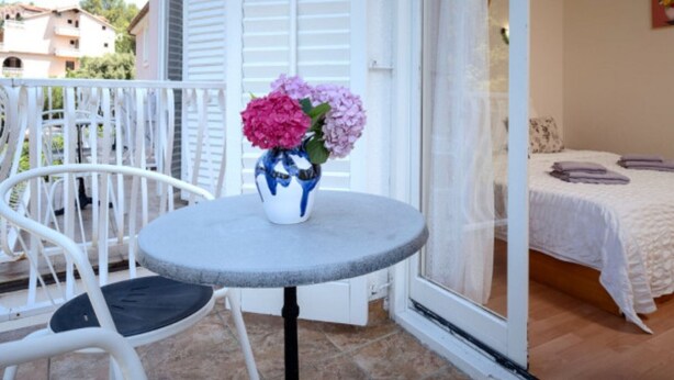 Een balkon met een witte stoel, een klein rond tafeltje met een bloevas en een bed met een witte spreidek.