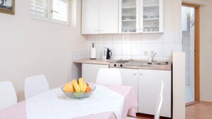 Een kleine keuken met witte kasten, een spoelbak en een fornuis. Er staat een tafel met een fruitschaal en een witte tafelkleed.