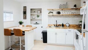Cozinha moderna com armários brancos, bancada de madeira e um espaço de bar com banquetas. Há um cesto de lixo e uma pequena prateleira com objetos decorativos.