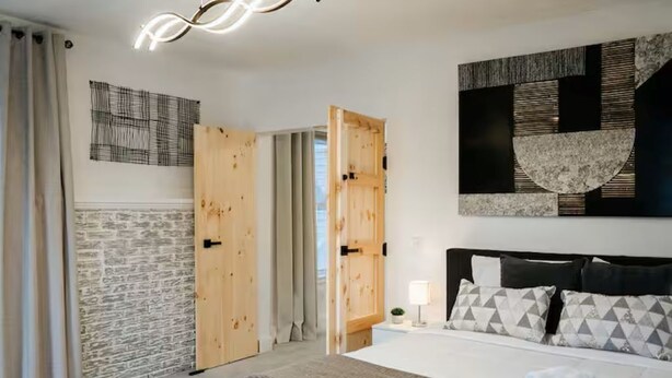 Quarto de hotel moderno com uma cama grande, porta de madeira, parede de pedra e uma obra de arte contemporânea sobre a cama.