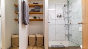 A bathroom with a glass shower enclosure, wooden shelves with towels, and two trash bins.