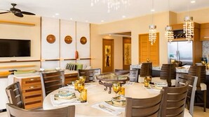 A modern dining area with a round table, chairs, and a chandelier. There is a television mounted on the wall and a ceiling fan.