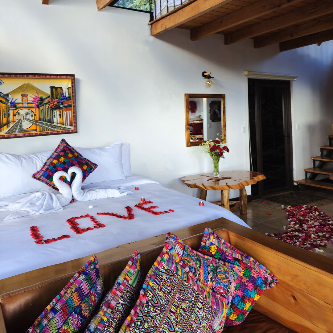 Un dormitorio con cama, almohadas coloridas, una decoración con forma de corazón sobre la cama, un cuadro en la pared, una mesita con un jarrón y una escalera.