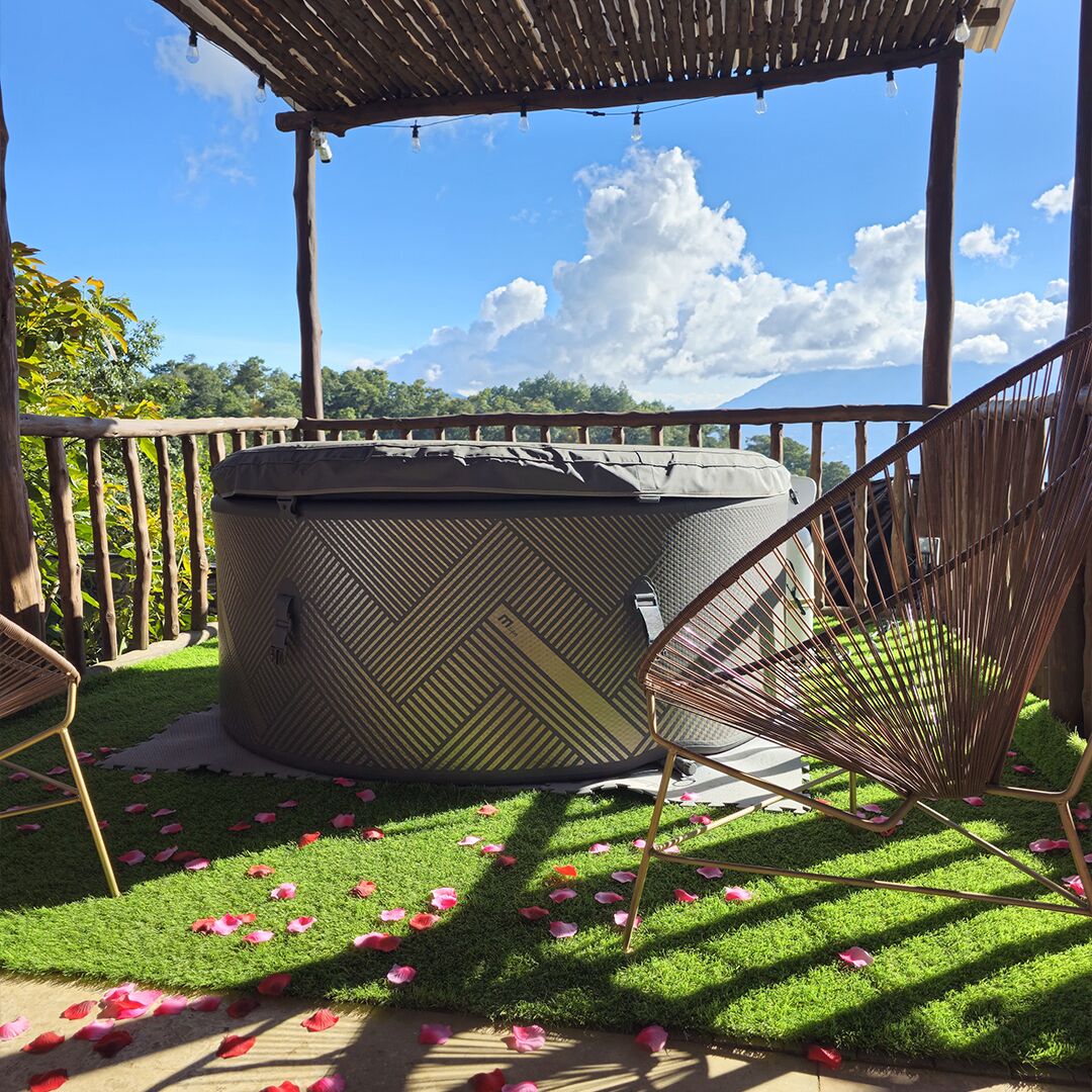 A hot tub under a wooden pergola with a hammock and chairs, surrounded by artificial grass and scattered rose petals.