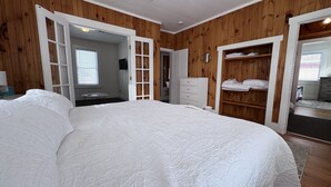 A bedroom with a large bed, wooden walls, a wardrobe, and a bathroom visible through an open door. - Historic Island Pond Home on VAST Trail – 3BR, 2BA (Brighton)