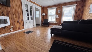 A living room with wooden floors, a dark sofa, a lamp, and a TV mounted on the wall. - Historic Island Pond Home on VAST Trail – 3BR, 2BA (Brighton)