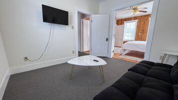 Une pièce avec un téléviseur blanc fixé au mur, une petite table blanche aux pieds dorés, un canapé noir et, au fond, un lit avec une tête de lit en bois.