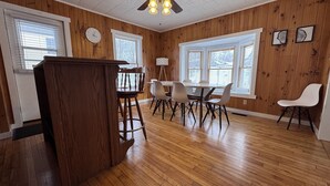 A dining area with a wooden table and chairs, a clock on the wall, and large windows with blinds. - Historic Island Pond Home on VAST Trail – 3BR, 2BA (Brighton)