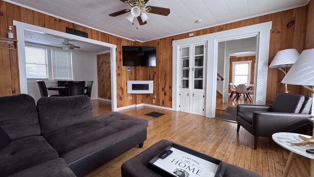 A modern living room with a grey sofa, a coffee table with a magazine, a TV mounted on the wall, and a wooden floor. - Historic Island Pond Home on VAST Trail – 3BR, 2BA (Brighton)