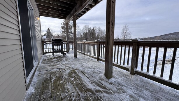 A covered porch with a wooden railing, a grill, and a view of a snowy landscape. - Island Pond Apartment on VAST Trail – 3BR, 2BA (Brighton)