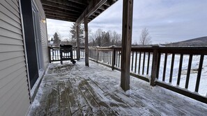 A covered porch with a wooden railing, a grill, and a view of a snowy landscape. - Island Pond Apartment on VAST Trail – 3BR, 2BA (Brighton)