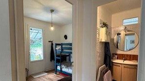 Een kamer met een houten vloer, een stapelbed, een klein tafeltje, een deur naar een andere kamer, en een badkamer met spiegel en wastafel.