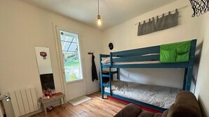 Een kleine, goed verlichte kamer met een stapelbed, een slaapbank, een radiator, een klein tafeltje en een raam met gordijnen.