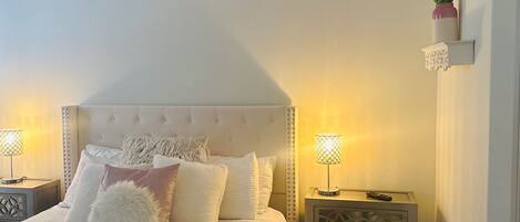 A bedroom with a large bed, two bedside tables with lamps, a ceiling fan, and a wall-mounted shelf with flowers.
