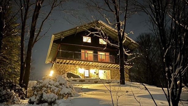 A snow-covered landscape with a wooden house featuring a balcony and illuminated windows. - Repère Öra | Spa, ski & nature (Saint-Adolphe-d'Howard)