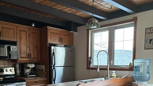A kitchen with wooden cabinets, a stainless steel refrigerator, a water dispenser, and a window with a view. - Repère Öra | Spa, ski & nature (Saint-Adolphe-d'Howard)