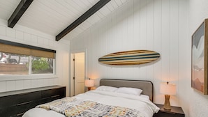 Quarto com uma cama, duas mesinhas de cabeceira com luminárias, uma janela, um prancha de surf fixada na parede e um quadro emoldurado.
