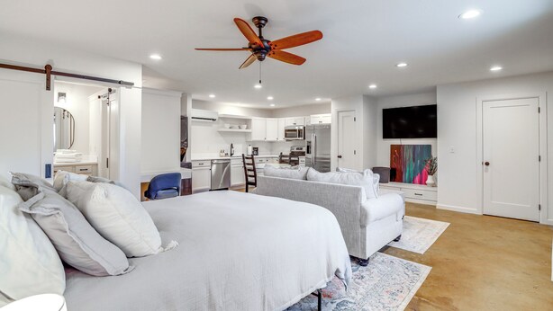 A bedroom with a large bed, two bedside tables, a ceiling fan, and a view into a kitchen area. - Central & Modern Studio w/ Workspace in Owasso! (Owasso)