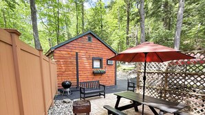 A wooden deck with picnic tables and an umbrella, surrounded by a wooden fence and a small cabin.
