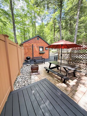 Eine Holzterrasse mit Picknicktischen und Sonnenschirm, eine kleine Holzhütte und ein Holzzaun.