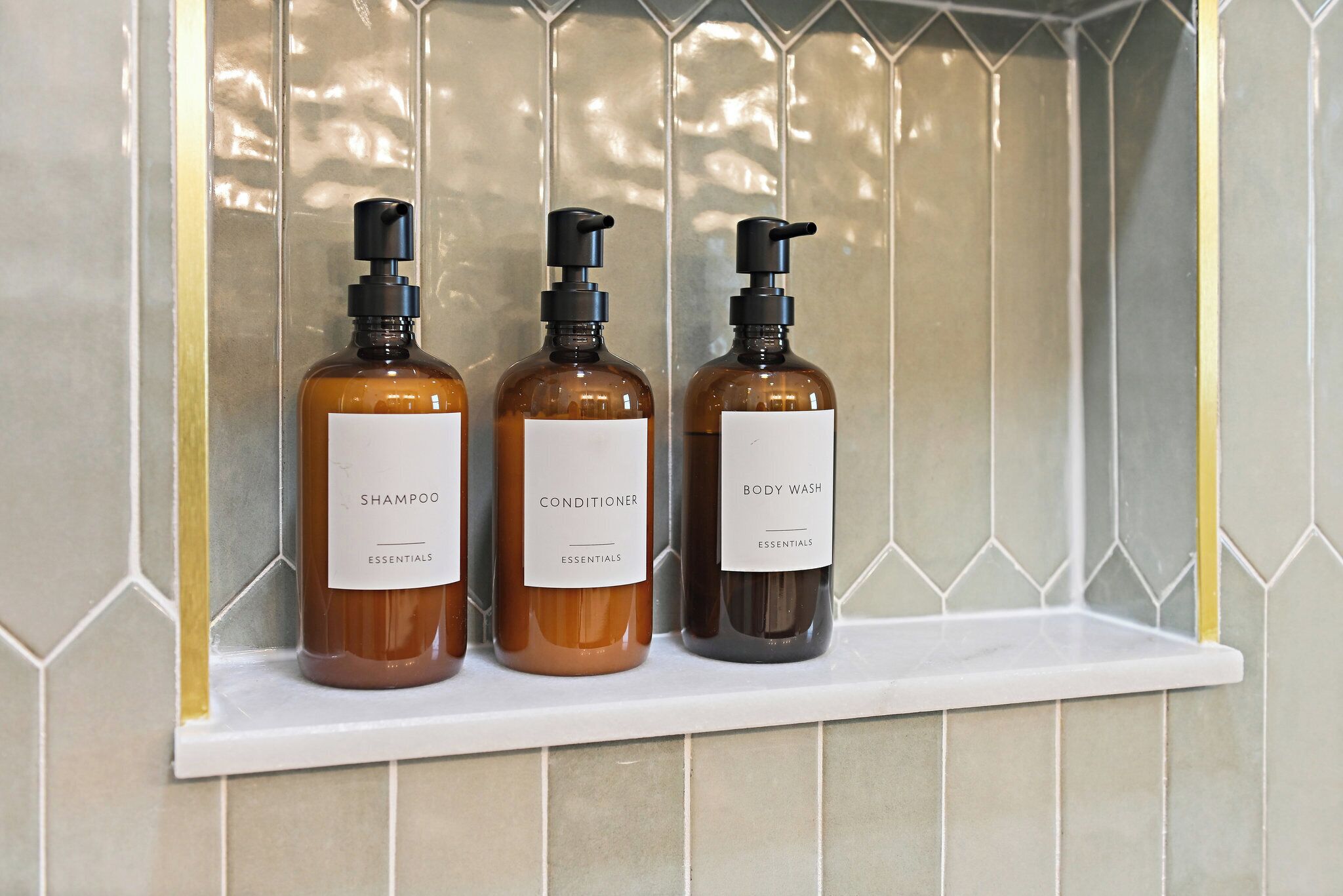 Drei Flaschen Shampoo, Conditioner und Duschgel stehen auf einem weißen Regal vor einer gefliesten Wand.