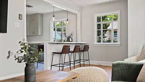 A modern kitchen with white cabinets, a central island, and hanging pendant lights. There are three bar stools and a round woven ottoman in the dining area.