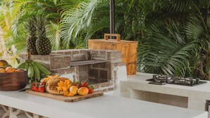 An outdoor kitchen with a stone wall, a hanging wicker light, and a wooden counter with stools. - The Sunset Shack (Ceiba)