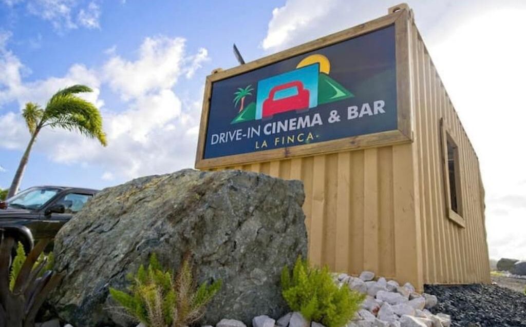 Estructura de cine al aire libre y bar, con una gran roca y palmeras en primer plano.