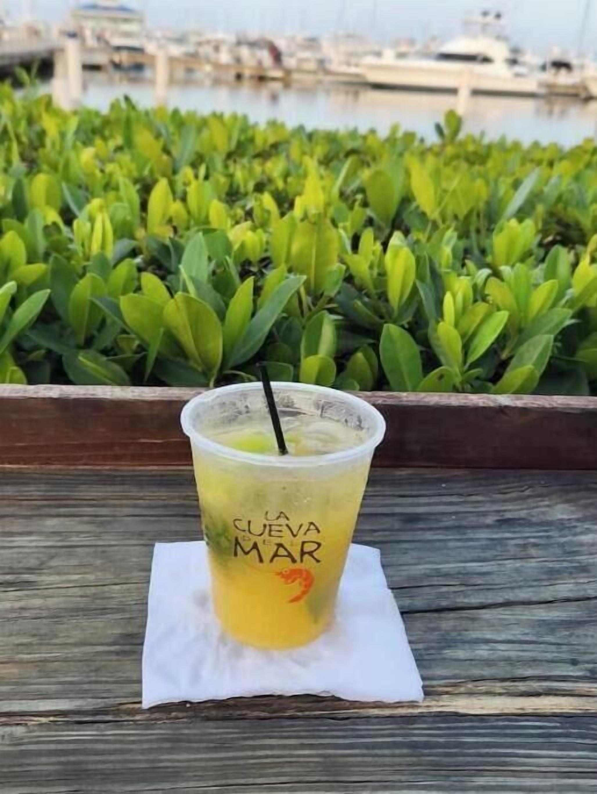 A plastic cup with a straw on a wooden table, labeled "LA CUEVA DEL MAR".