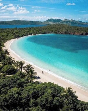 Ein Strand mit klarem türkisfarbenem Wasser, umgeben von üppiger, grüner Vegetation und Hügeln im Hintergrund.
