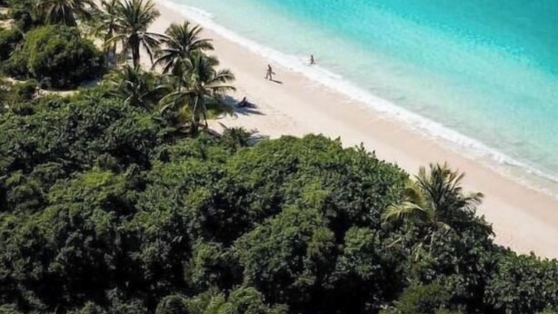 Luchtbeeld van een strand met helder turquoise water, omgeven door weelderige groene begroeiing en heuvels op de achtergrond.
