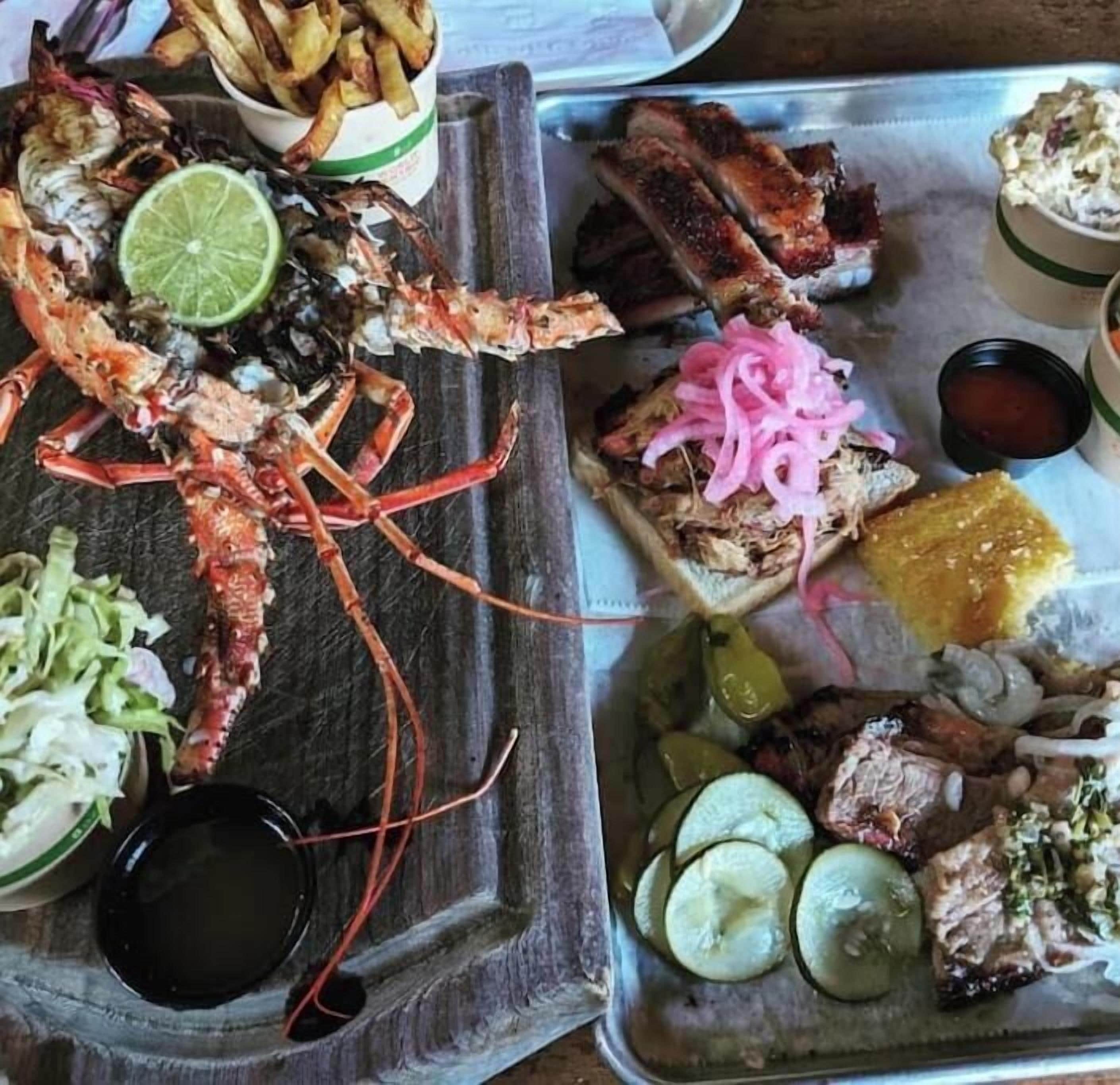 Un plateau de fruits de mer comprenant du homard, des travers de porc et divers accompagnements, notamment de la salade de chou et des cornichons.