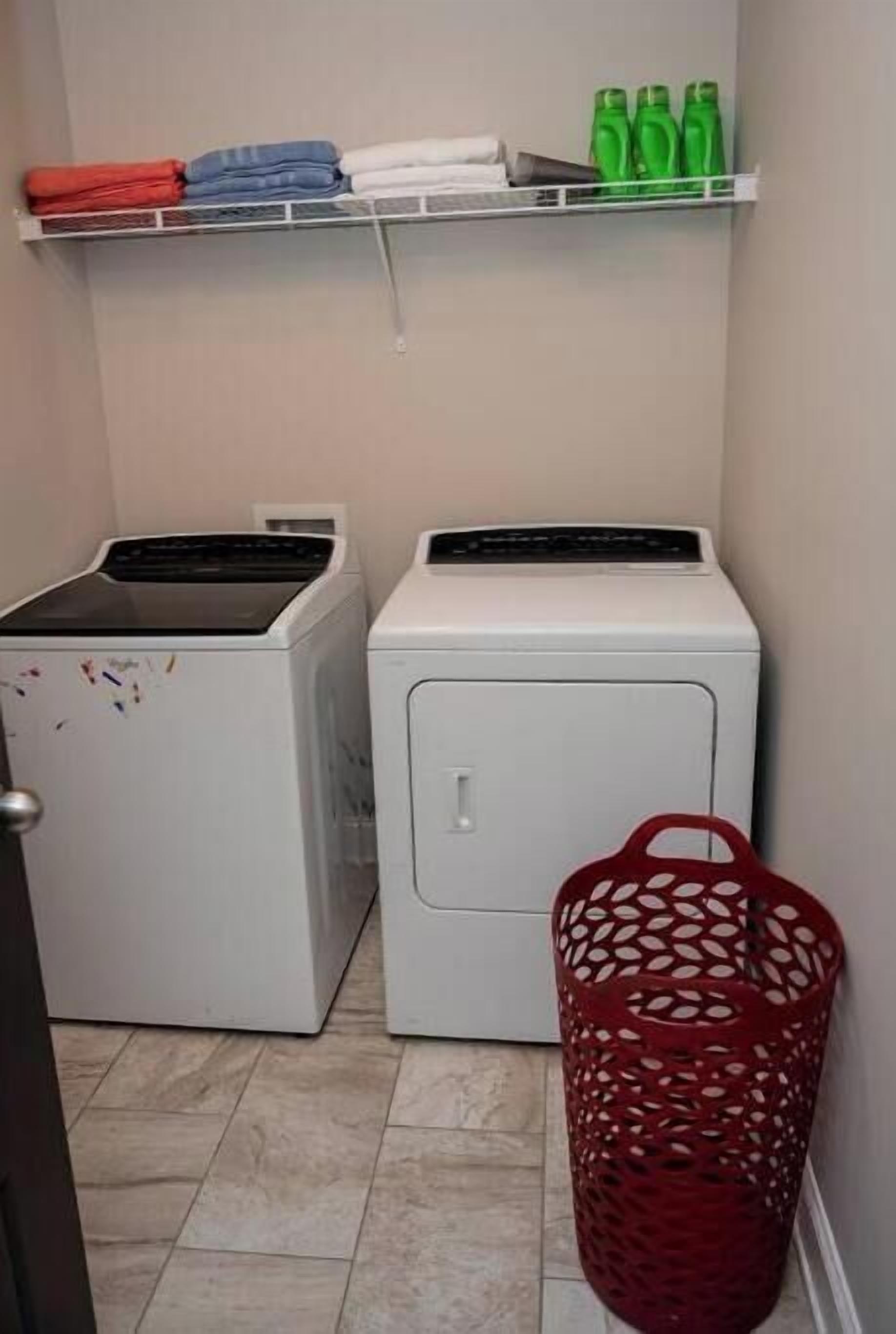 Een wasruimte met twee wasmachines, een plank met opgevouwen kleding en groene flessen, en een rode wasmand.