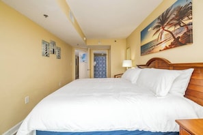 A hotel room with a large bed, a wooden headboard, a painting of a beach scene, and a door leading to another room.