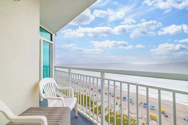 A balcony with white chairs and a view of the beach with umbrellas.