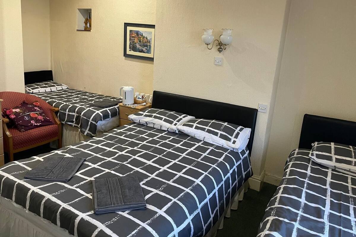 A hotel room with three beds, each with a black and white checkered bedspread. There is a chair with a floral cushion, a small table with a kettle and cups, and a framed picture on the wall.