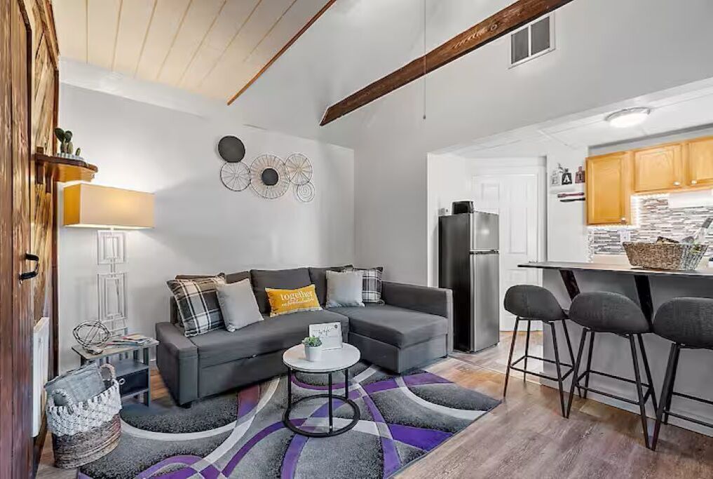 A modern living room with a grey sofa, a small round coffee table, and a kitchen area with a refrigerator and bar stools.