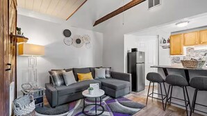 Una sala de estar moderna con un sofá gris, una mesita de centro redonda y una cocina con nevera y taburetes de bar.