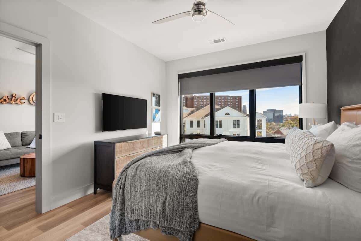 A bedroom with a large window, a bed, a TV, and a ceiling fan.