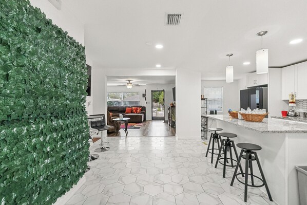A modern kitchen with a white marble floor, a green leafy wall, and a dining area with stools.