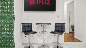 Ein Zimmer mit an der Wand montiertem Fernseher, der "NETFLIX" anzeigt, zwei schwarzen Barhocker und einer Wanddekoration mit grünen Blättern.
