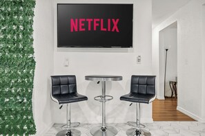 Una stanza con una TV a muro che mostra "NETFLIX", due sgabelli neri e una decorazione parietale con piante verdi.