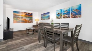 Ein Essbereich mit Holztisch und Stühlen, einem Fernseher links sowie drei an der Wand hängenden Bildern, die Meeresansichten zeigen.