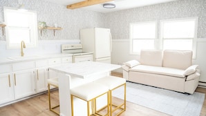 Une cuisine moderne avec des meubles blancs, un plan de travail blanc, un tabouret de bar à cadre doré, un canapé blanc et un pare-feu décoratif.
