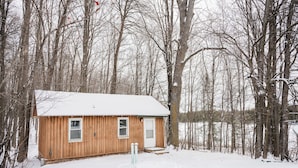 Une petite cabane en bois, dont le toit est recouvert de neige, entourée d’arbres et posée sur un sol enneigé.