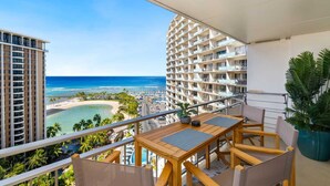 Ein Balkon mit Holztisch und -stühlen, der Blick auf einen Strand und das Meer mit einem Hochhaus im Hintergrund freigibt.