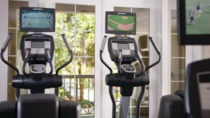 Two exercise bikes with mounted screens displaying sports games, in a room with large windows and a view of greenery outside. - Marriott’s Harbour Club 2-Bedroom Villas – The Perfect Lowcountry Getaway (Hilton Head Island)