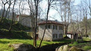 A stone house with a red-tiled roof, surrounded by trees and a small stream.